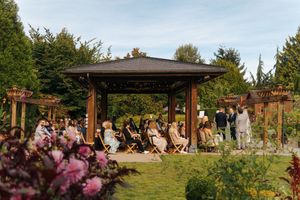 Outdoor wedding ceremony in a lush garden with guests seated under an elegant wooden pavilion on a sunny day.