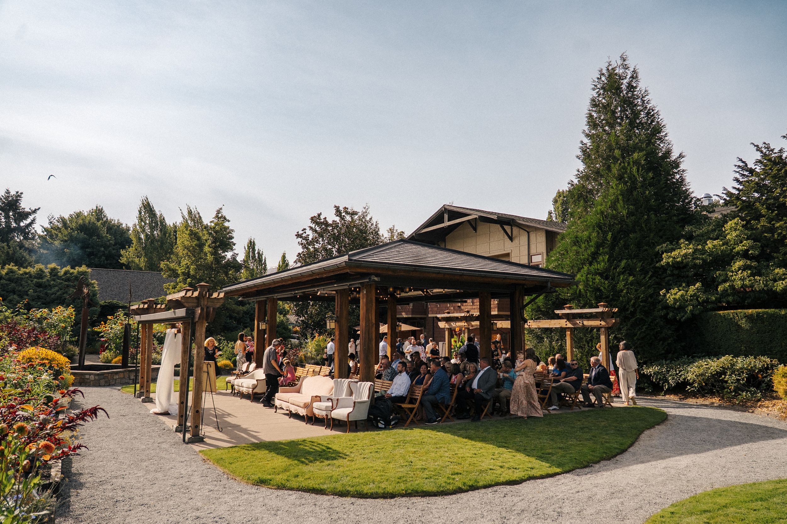 Outdoor wedding ceremony under a wooden pavilion surrounded by lush gardens and tall trees on a sunny day.