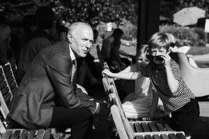Two people conversing outdoors on wooden chairs in black and white vintage photograph.