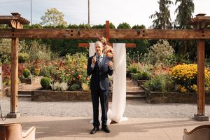 Man in blue suit speaks at outdoor garden wedding ceremony with colorful flowers and wooden pergola backdrop.