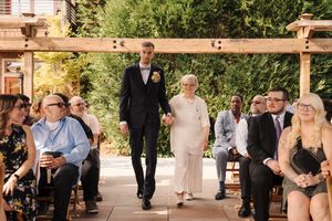 Groom in navy tuxedo walks elderly woman down outdoor wedding aisle lined with seated guests under wooden pergola.