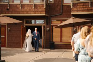 Bride in white gown and veil walks with escort through venue doors at outdoor wedding ceremony.