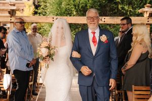 Bride in white gown and veil walks down aisle with older man in navy suit and red tie at outdoor wedding ceremony.