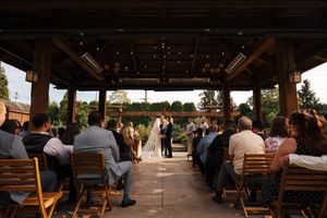 Outdoor wedding ceremony under a rustic wooden pavilion with string lights, guests seated in wooden chairs facing the couple.