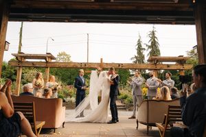 Bride and groom share a kiss at an outdoor wedding ceremony with guests seated in a rustic venue surrounded by greenery.