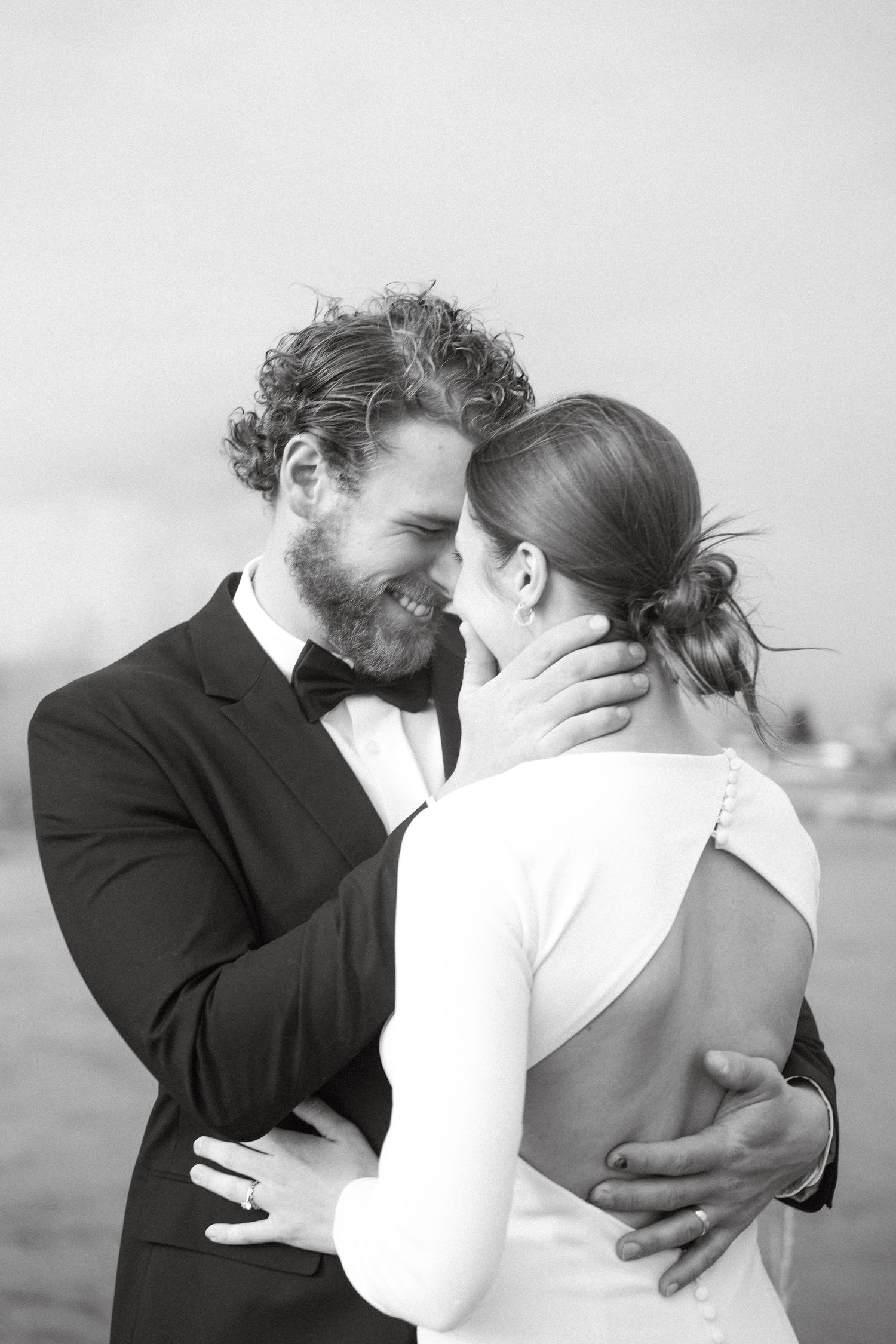 Black and white photo of a smiling groom in a tuxedo embracing his bride in a white wedding dress outdoors.