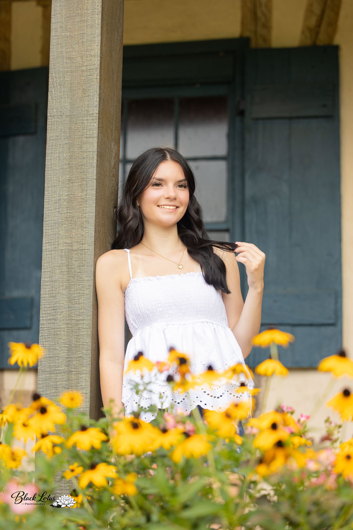 Young woman in white sundress smiling among yellow black-eyed Susan flowers by a rustic porch column.