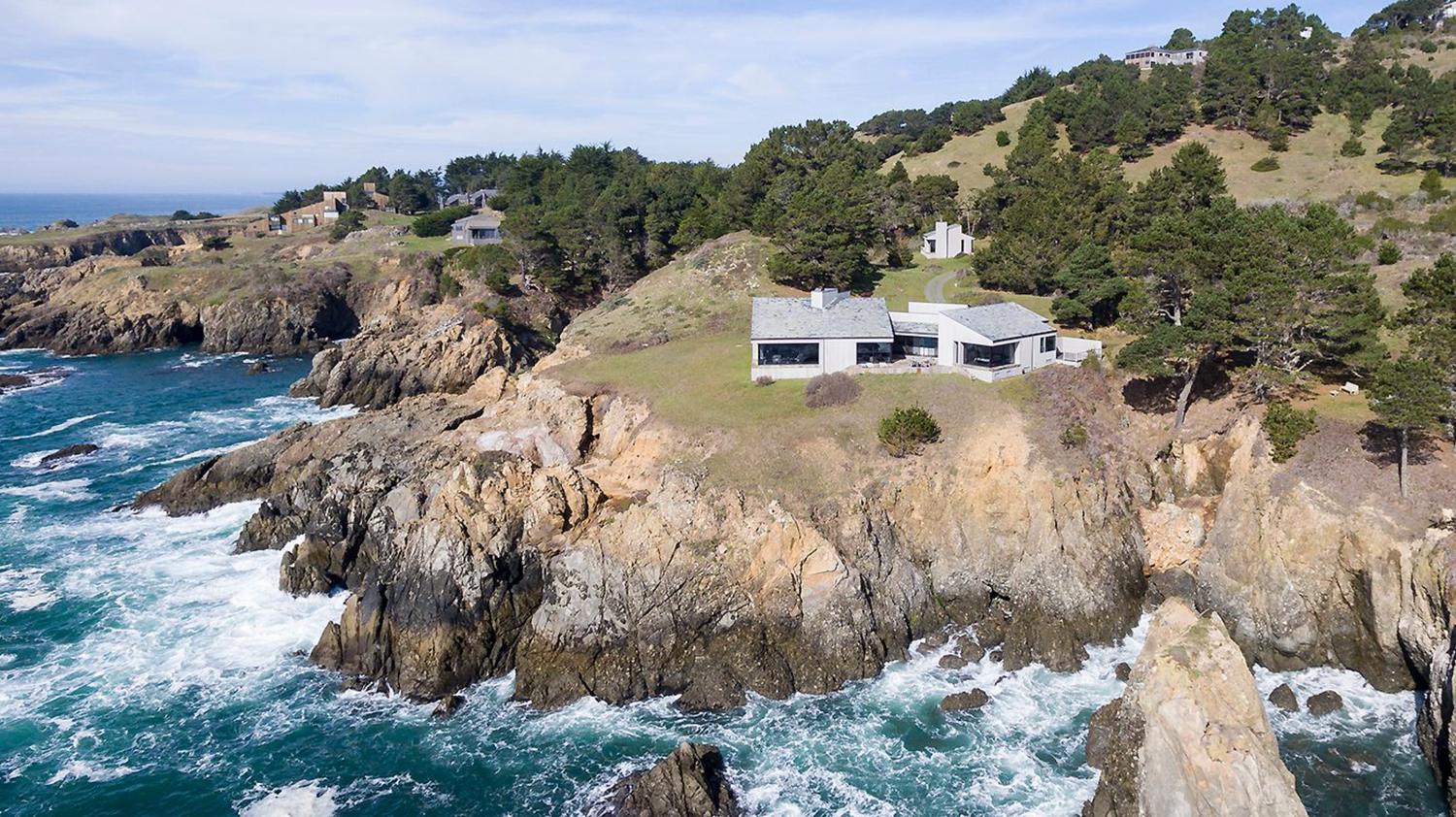 Aerial view of modern buildings perched on dramatic rocky cliffs overlooking turbulent blue ocean waters.