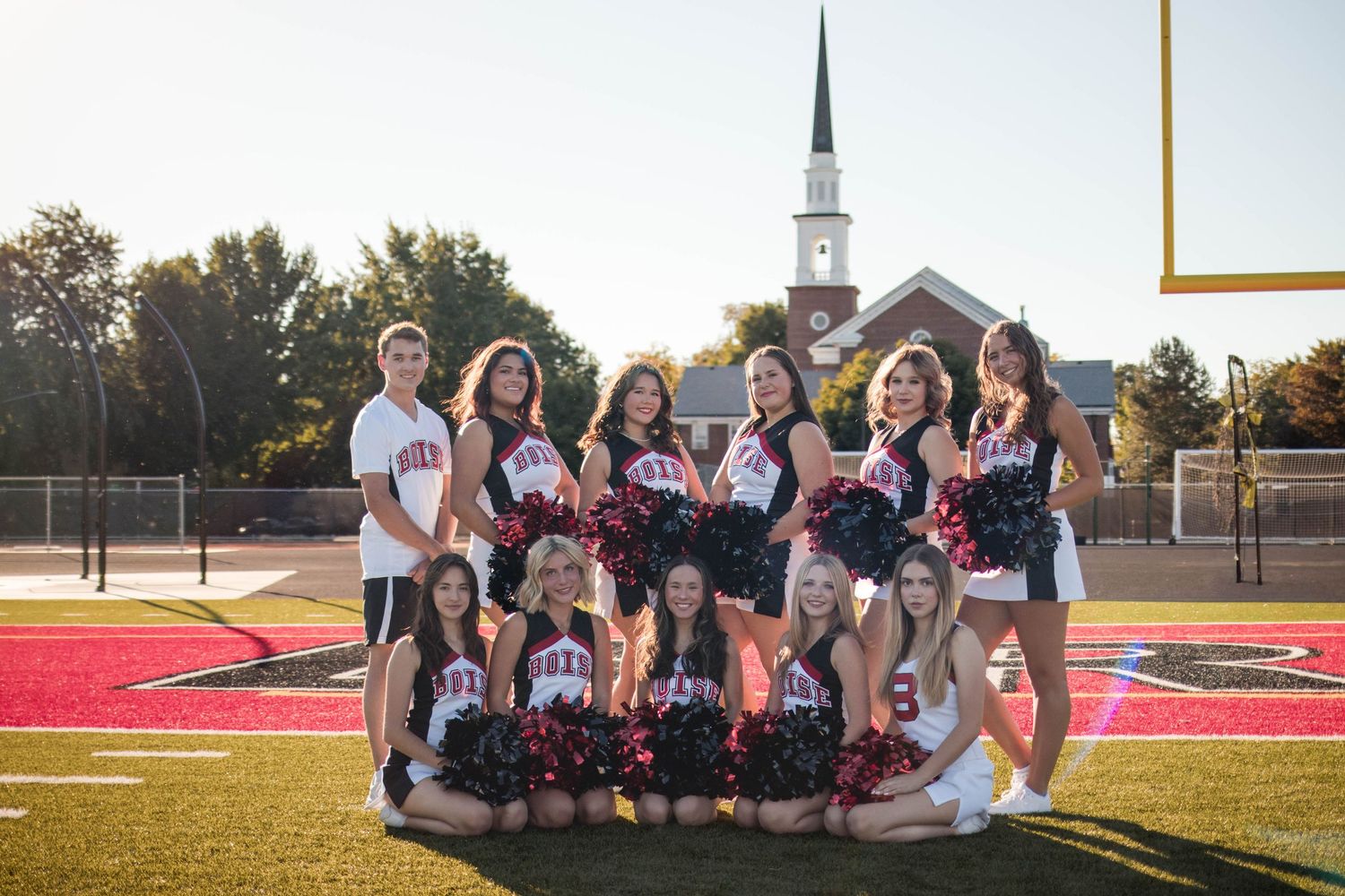 High school cheerleading squad posing on football field in red and white uniforms with pom-poms on a sunny day.