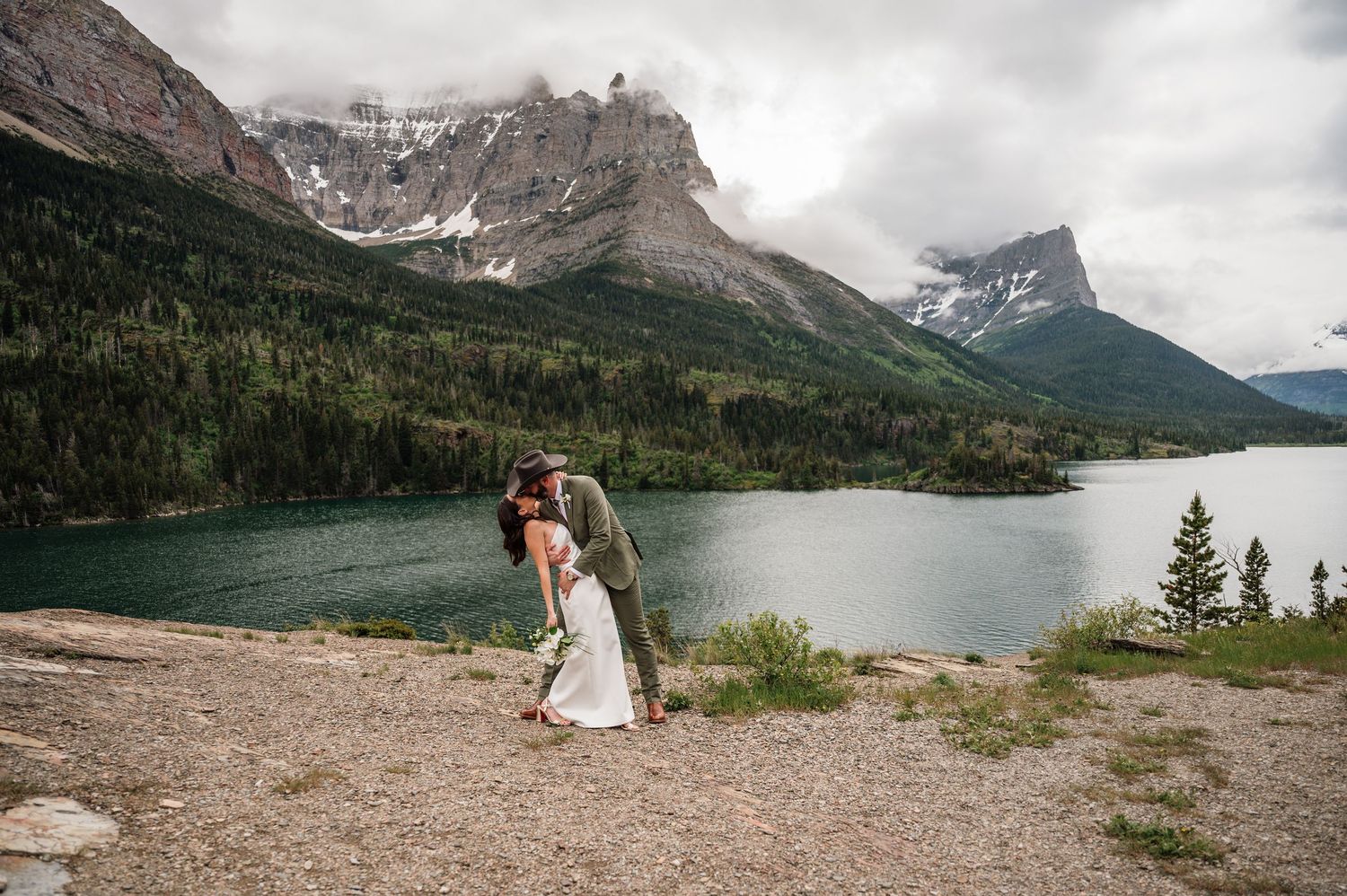 Bride and groom share a romantic dip kiss on a rocky overlook with a turquoise mountain lake and peaks behind them.