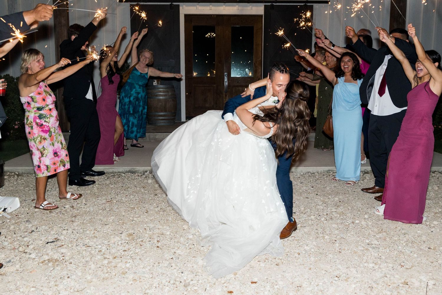 Groom dips bride during sparkler exit at outdoor wedding reception with guests lining the path.