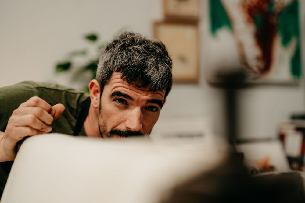 Man peering closely over the edge of a printing press roller, carefully inspecting his work in an art studio.