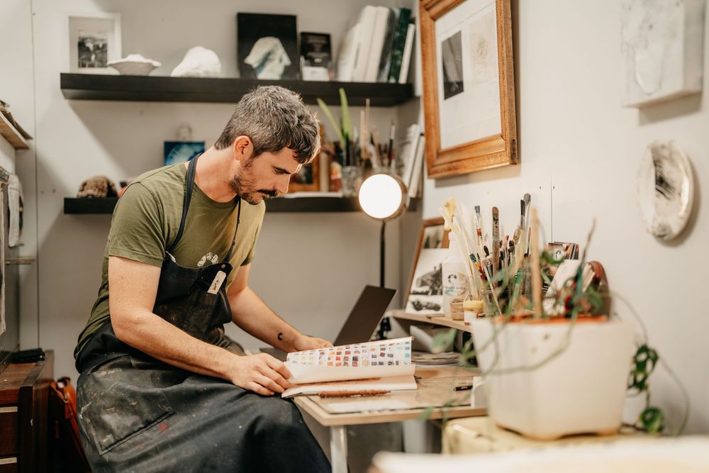 A craftsman in a green shirt and apron works at a wooden desk in an art studio.