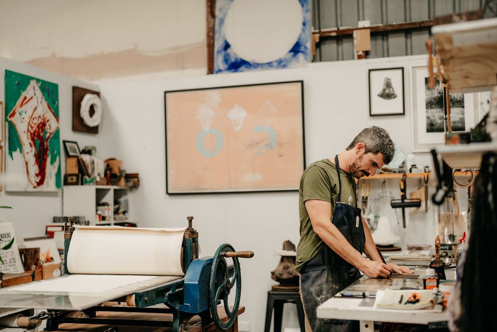 Artist in an apron working at a printing press in a bright studio filled with artwork and creative tools.