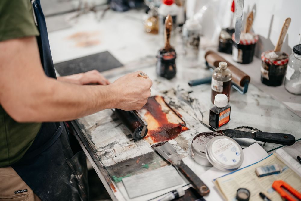 Artist applying heat to a colorful block print in a cluttered printmaking studio.