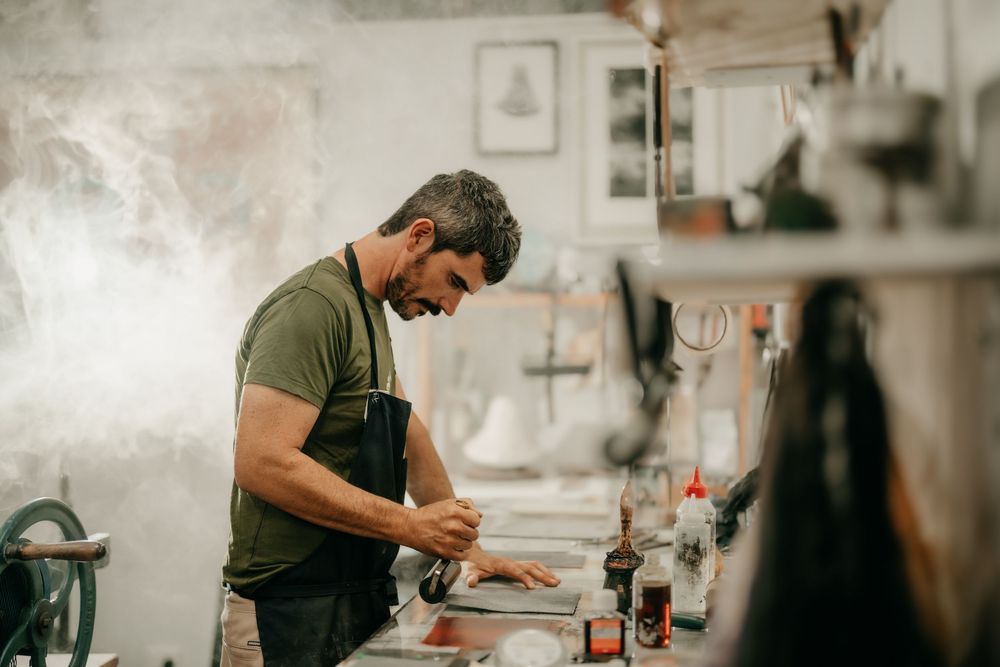 A focused male artist in an apron works at a printmaking bench surrounded by inks.