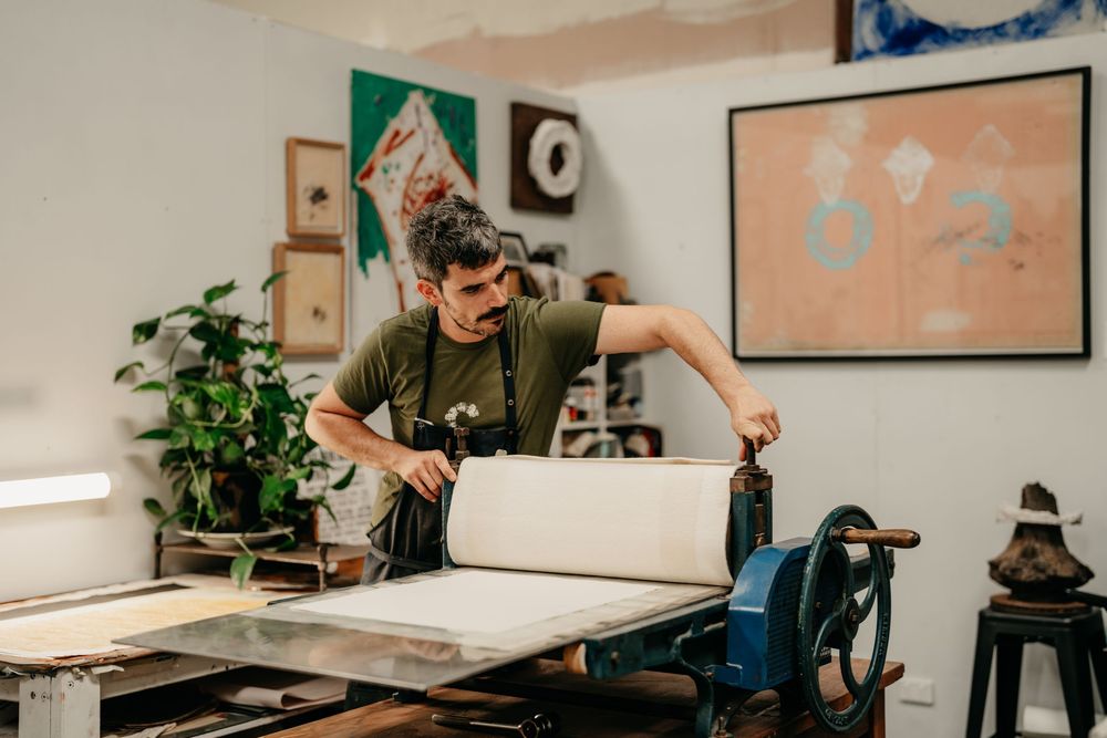 Man in a green shirt operating a large printing press roller in an art studio with colorful paintings on the wall.