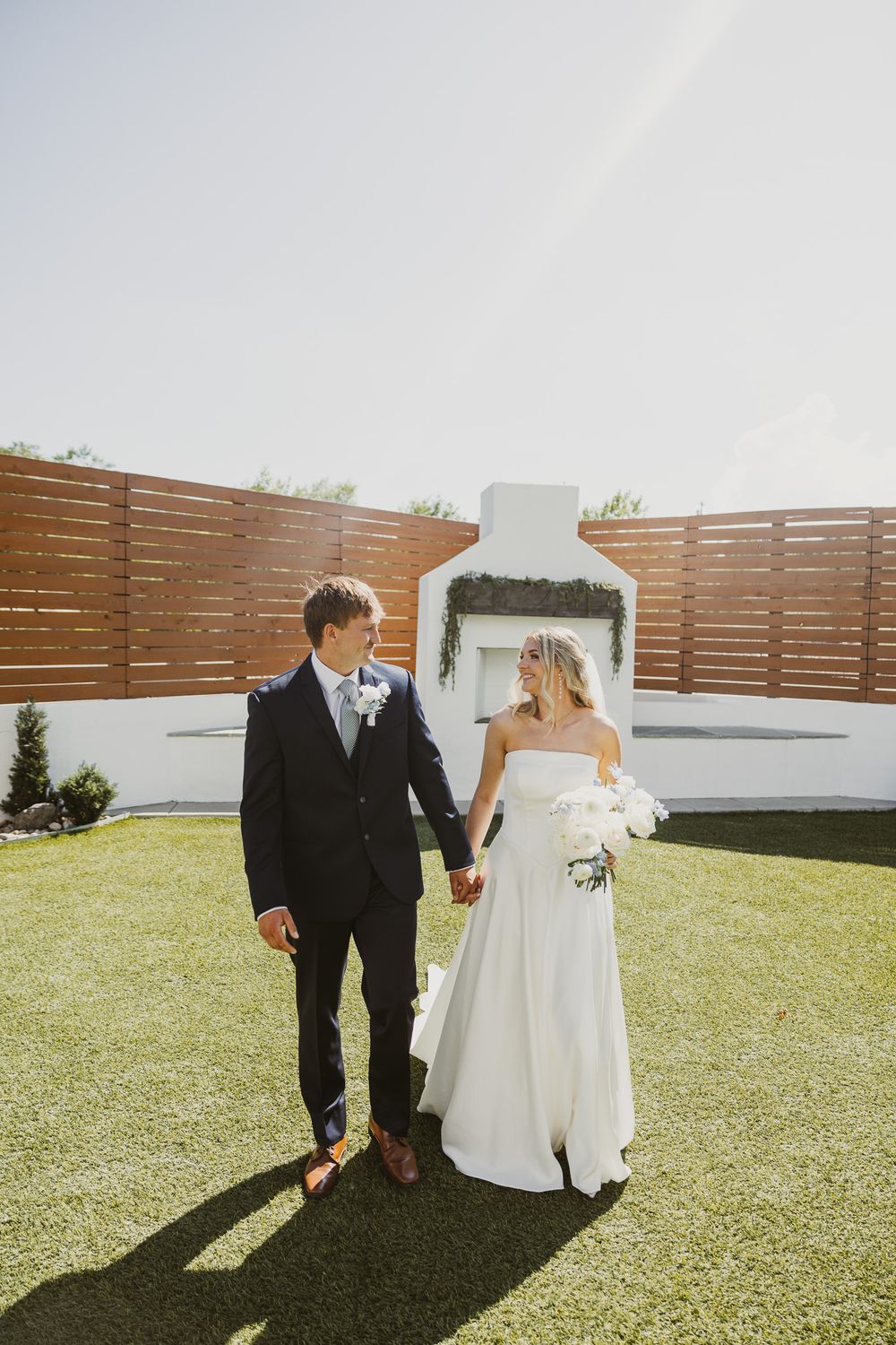 A bride and groom hold hands walking on green grass in front of a white outdoor fireplace on their wedding day.