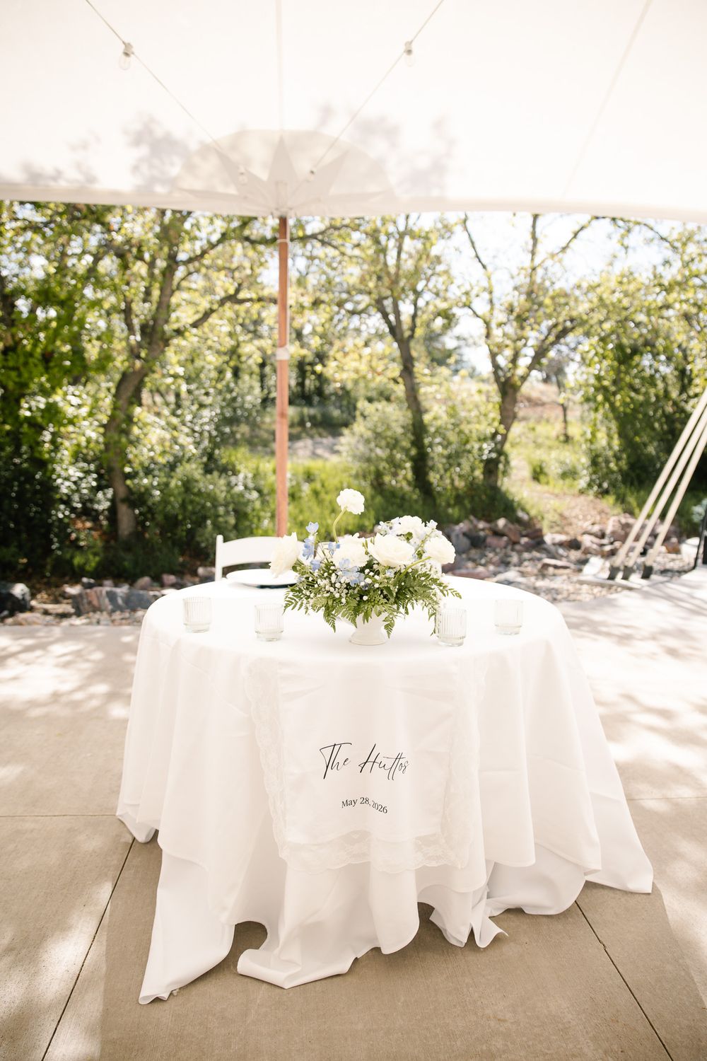 Elegant wedding sweetheart table with white linen, floral centerpiece, and personalized tablecloth under a tent.