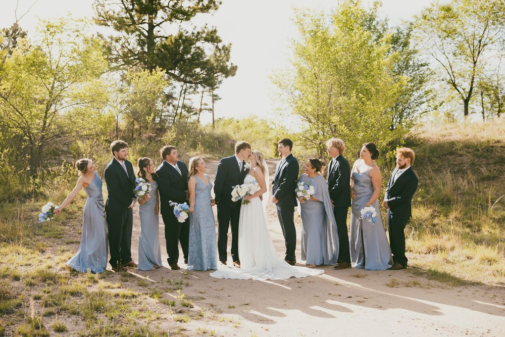 Wedding party poses outdoors in sandy setting, bridesmaids in blue-gray gowns flanking bride and groom.