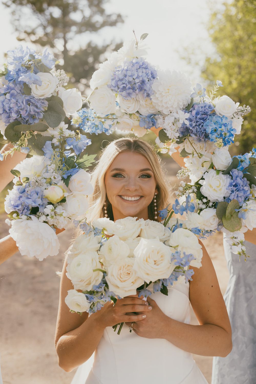 Smiling bride holding white rose bouquet framed by blue hydrangea and white floral arrangements outdoors.