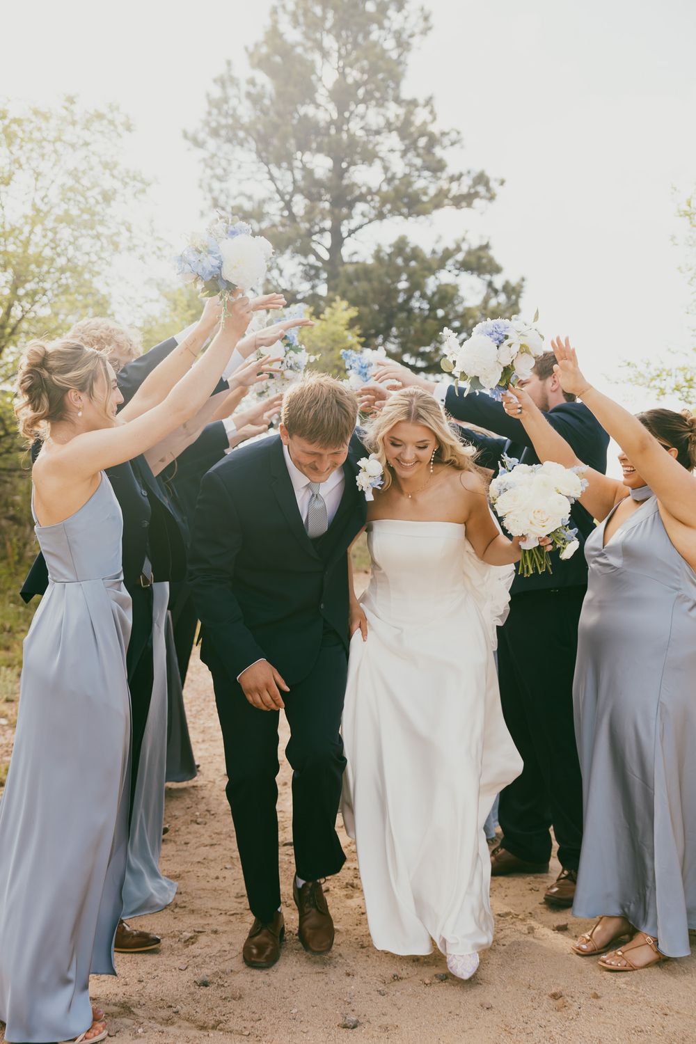 Bride and groom walk through floral arch tunnel created by wedding party in dusty blue dresses outdoors.