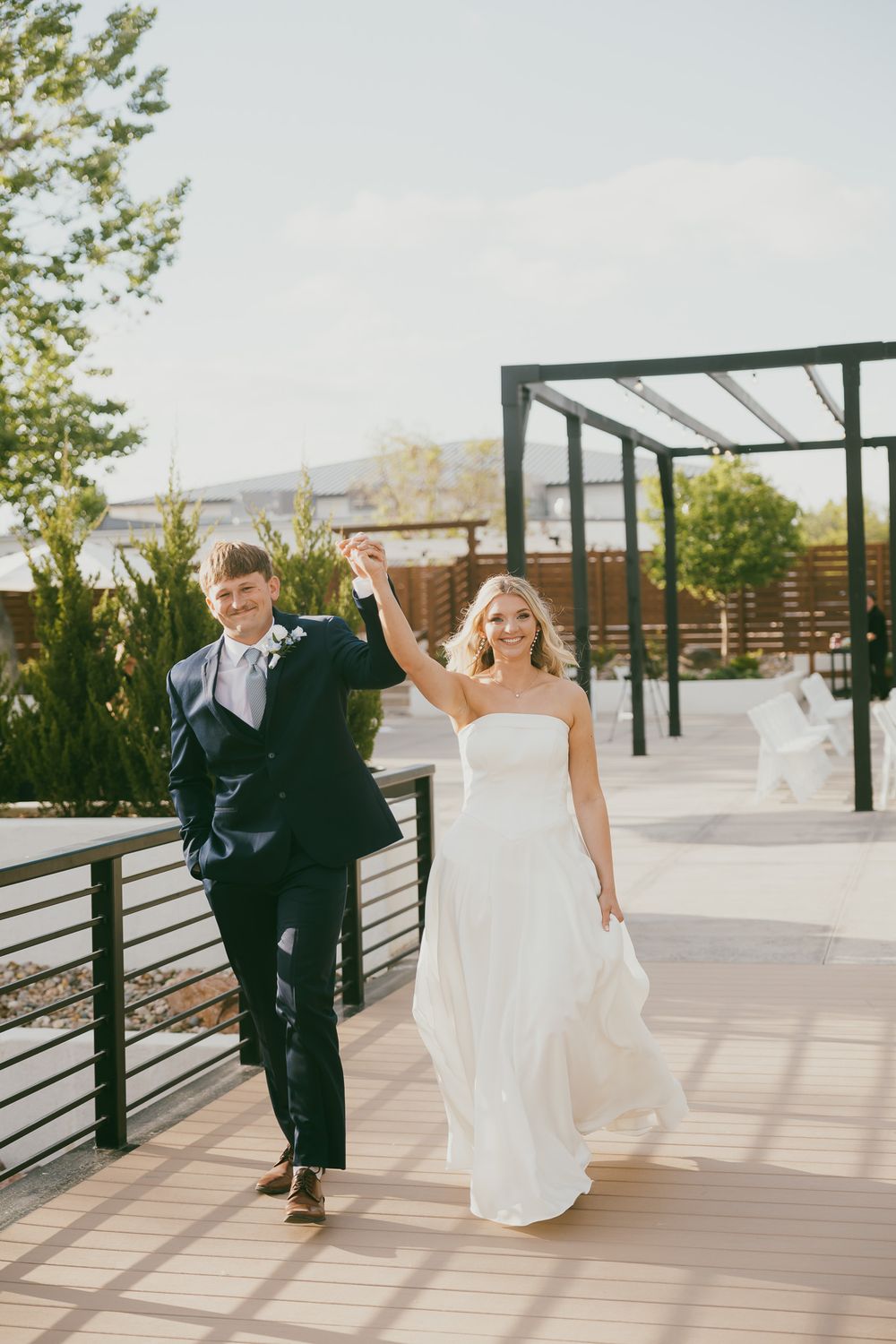 A joyful bride and groom walk hand-in-hand raised on a sunny outdoor deck, celebrating their wedding day.