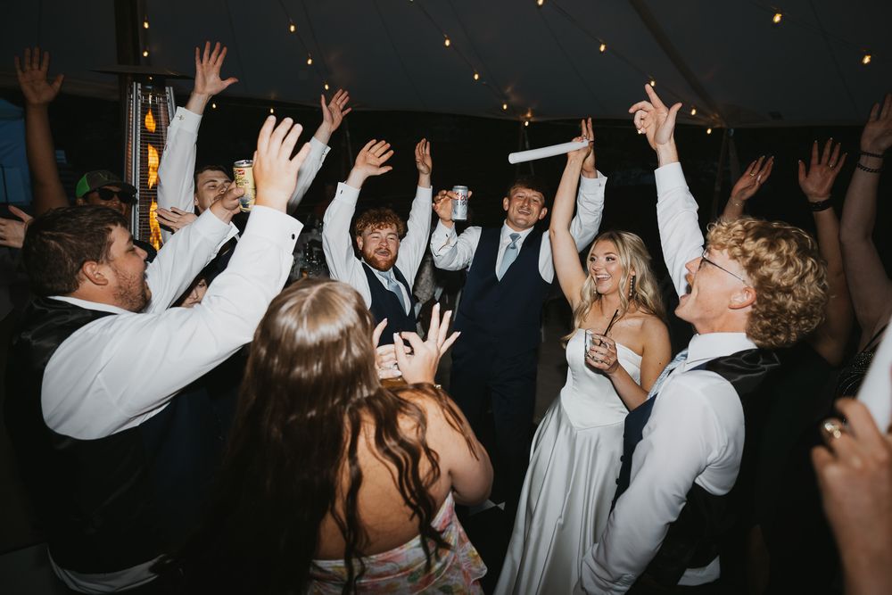Wedding guests celebrate on the dance floor with hands raised at a festive indoor reception under warm string lights.