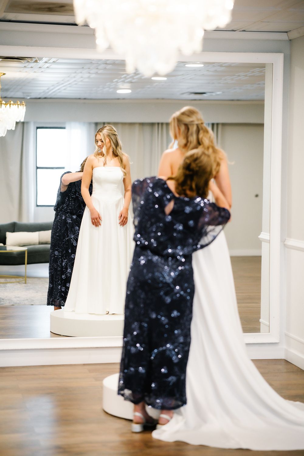 Bride tries on a white wedding gown while a woman in a navy sequin dress assists her in an elegant bridal boutique.