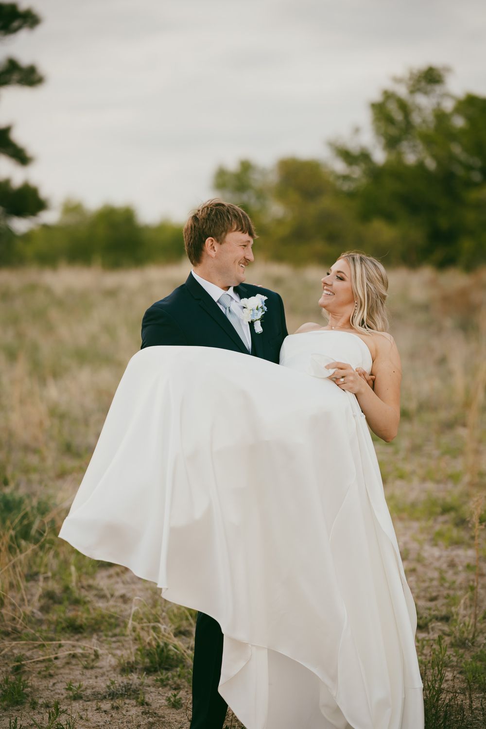 Groom in navy suit carries smiling bride in white strapless gown through a scenic outdoor field on their wedding day.