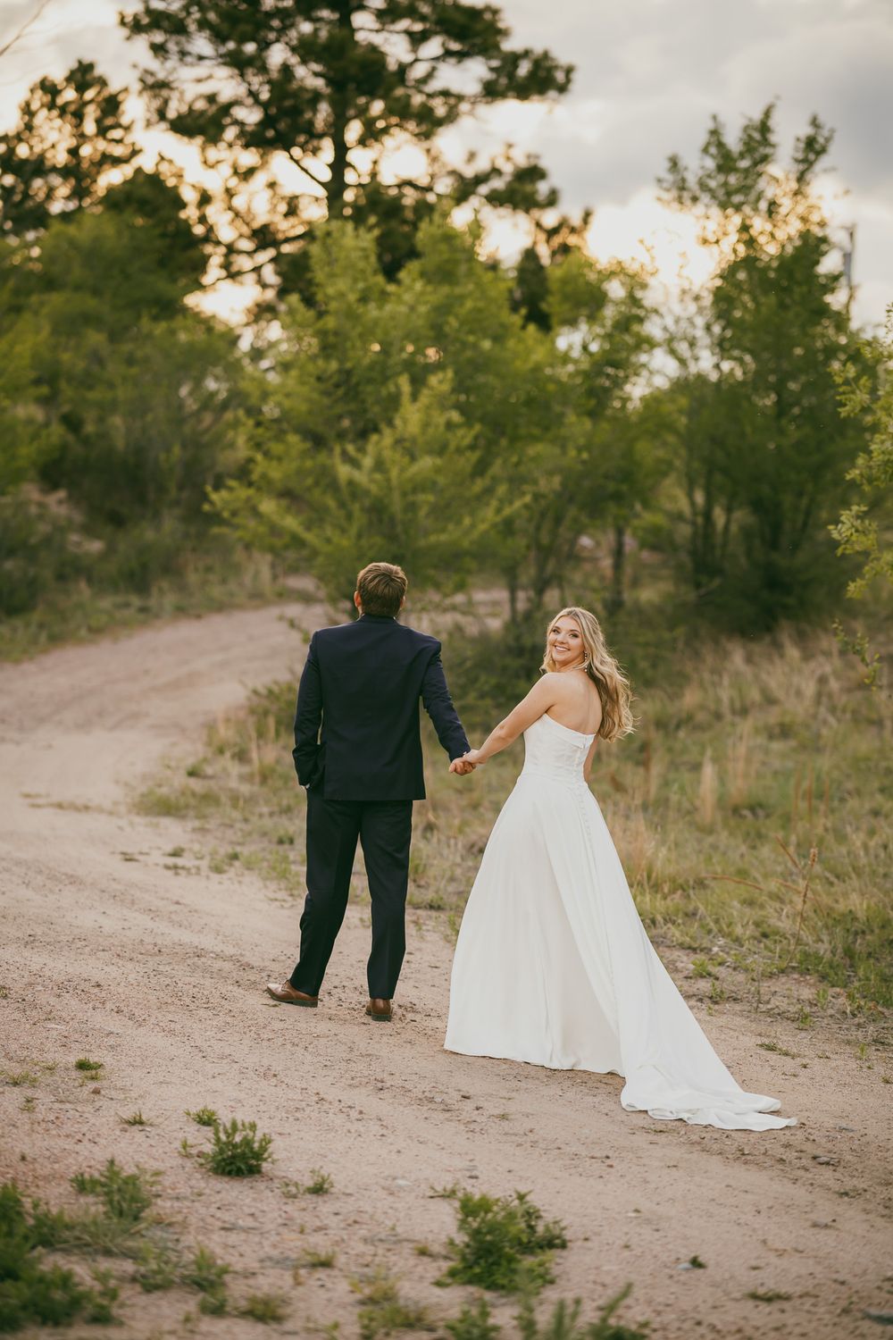 Bride in white gown holds groom's hand on a dirt path, glancing back with a smile, surrounded by lush green trees.