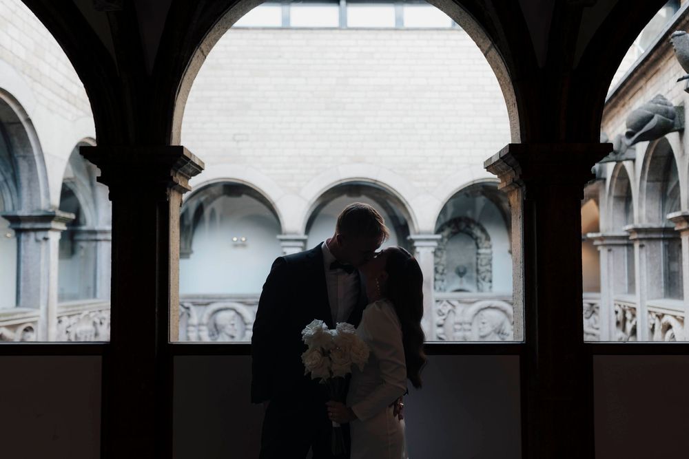 Silhouette of a couple kissing under elegant arched columns in a historic European courtyard, bride holding white bouquet.