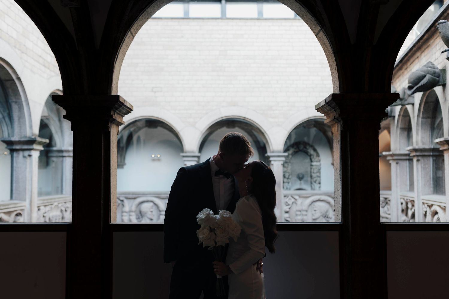Silhouette of a couple kissing under elegant arched columns in a historic European courtyard, bride holding white bouquet.