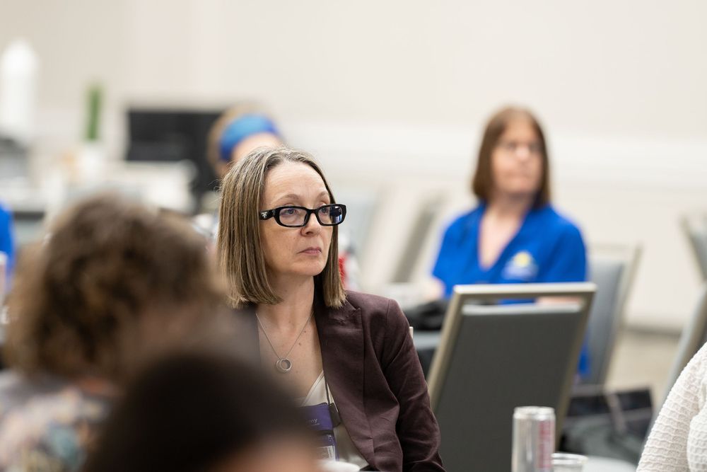 Woman with glasses and brown blazer attentively listening in a professional meeting or classroom setting.