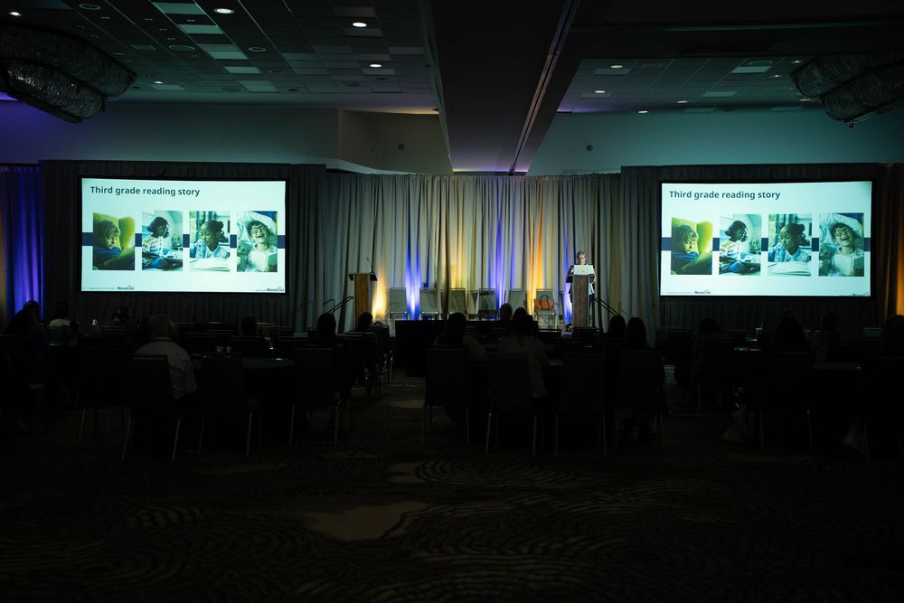 A dimly lit conference hall with attendees seated at tables facing two large projection screens displaying a third-grade reading story.