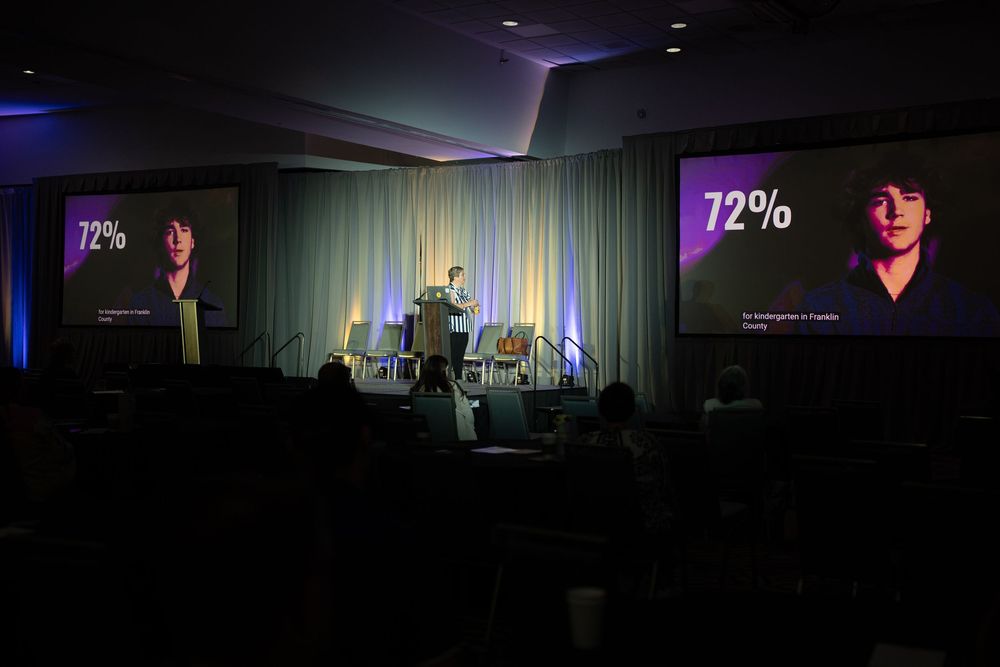Speaker presenting on stage at a conference with large screens displaying 72% statistic and a portrait photo in a dark auditorium.