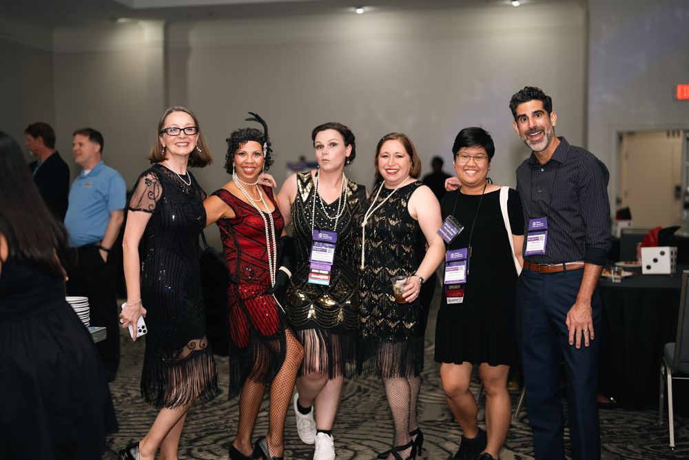 Group of six people posing together at a formal gala event, wearing elegant cocktail attire and conference lanyards.