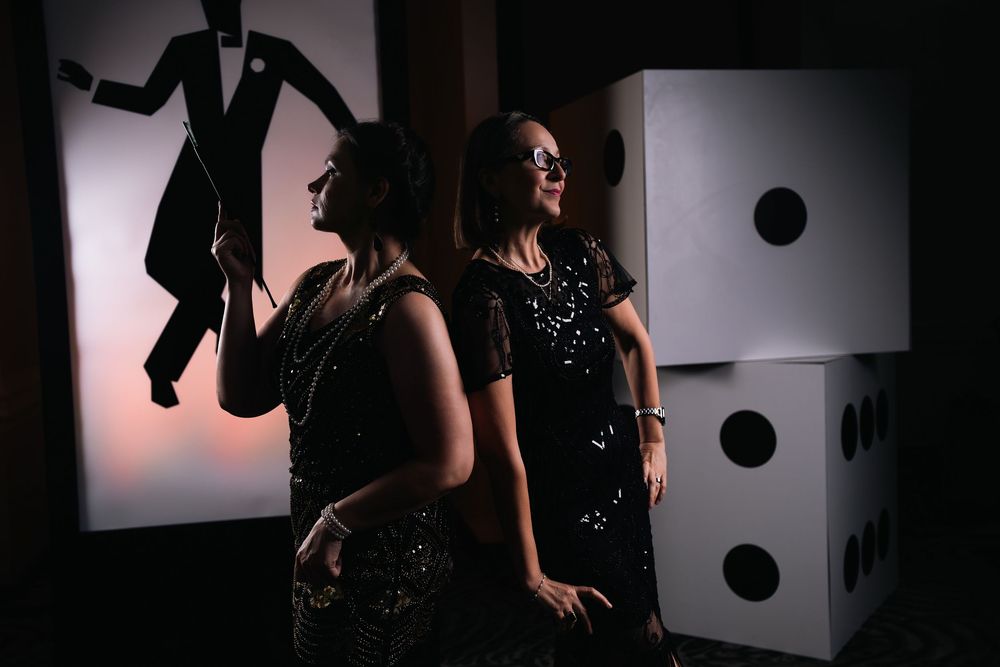 Two women in black sequin dresses pose near oversized dice and a James Bond silhouette at a casino-themed event.