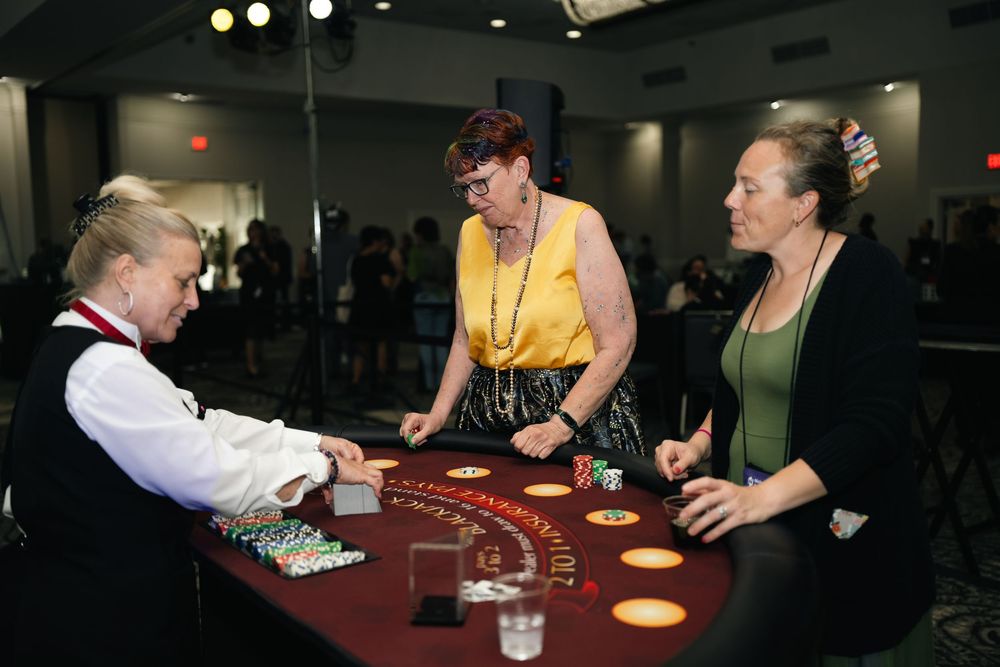 Two women play blackjack at a casino table with a dealer in a formal vest at an indoor event.