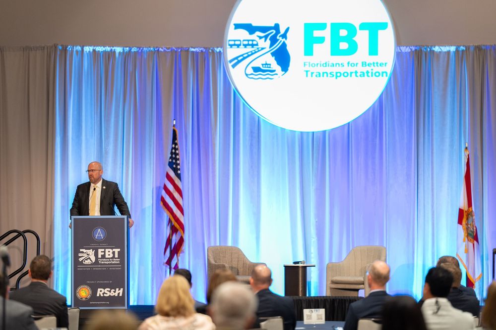 Speaker at FBT Floridians for Better Transportation conference podium with American and Florida state flags.