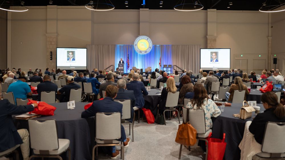 Large conference hall with attendees seated at round tables facing a stage with speakers and presentation screens.