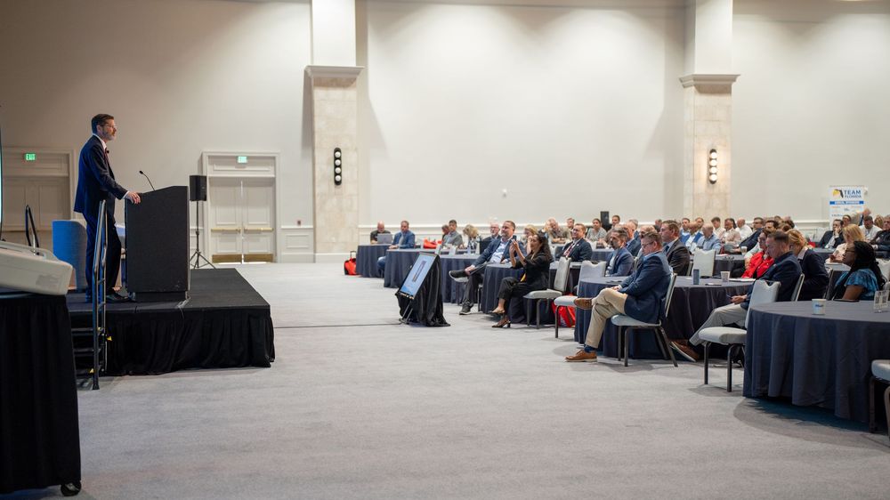 Speaker presenting at a professional conference in a large convention hall with an attentive seated audience.