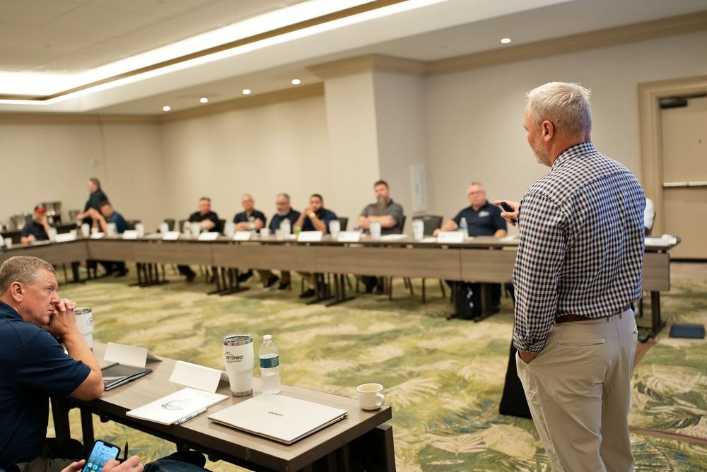 Business professionals attend a corporate meeting in a conference room with a speaker presenting at the front.