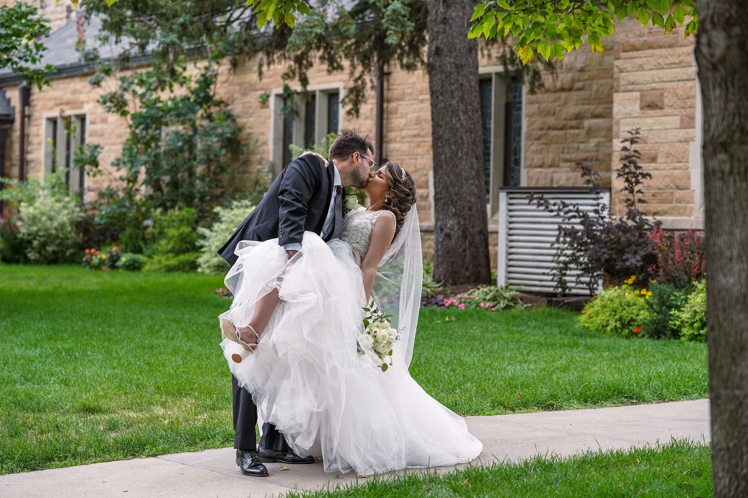 Groom dipping bride in elegant wedding dress on garden pathway beside stone building.