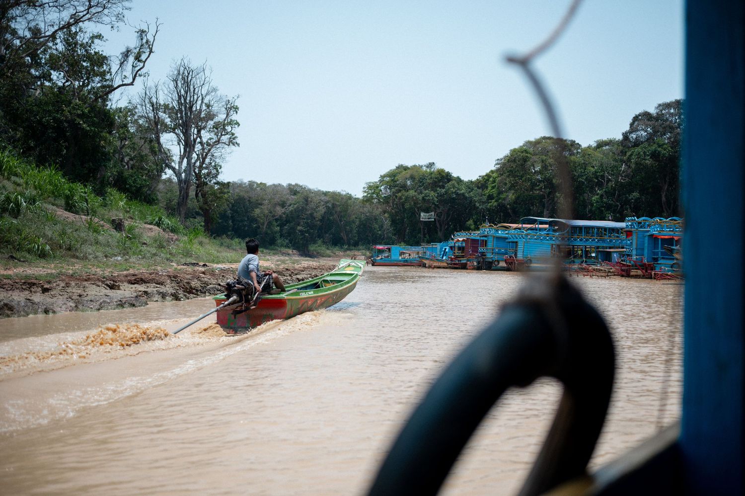 Boats navigating a muddy brown river surrounded by lush green trees in a tropical Southeast Asian waterway.