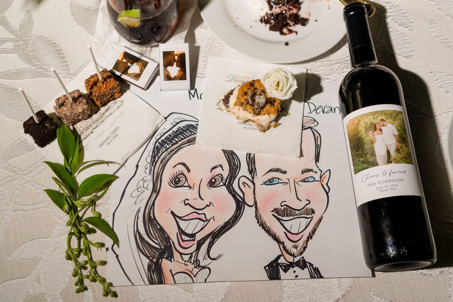 Caricature drawing of a bride and groom on a wedding table with a custom wine bottle, food, and decorative greenery.