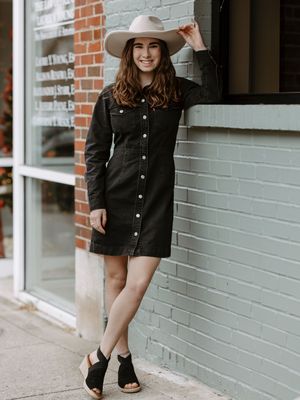 Woman in black denim button-up dress, wide-brim hat, and wedge booties leaning against a brick wall outdoors.