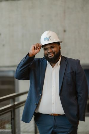 A smiling Black businessman in a navy suit tips his white construction hard hat while posing indoors.