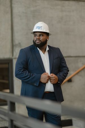 Professional Black man in navy suit and white hard hat stands confidently at a construction site.