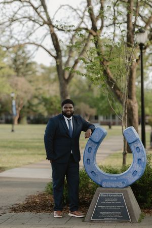 Man in navy suit posing next to Blue Horseshoe sculpture on college campus surrounded by trees.